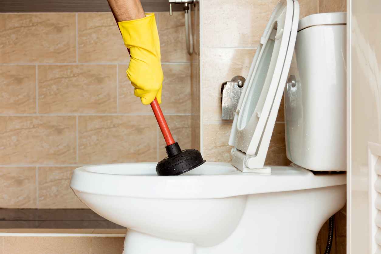  Human hand wearing yellow rubber gloves is using a plunger to fix a clogged toilet bowl. 