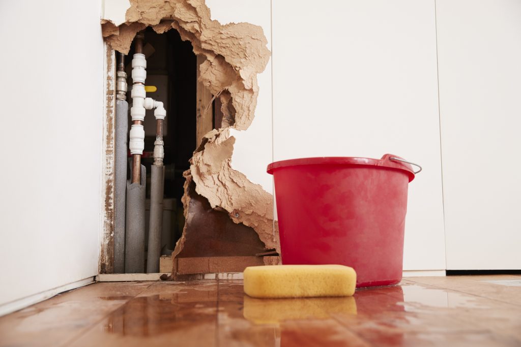 Damaged wall, exposed burst water pipes, sponge and red bucket on wet wood floor.